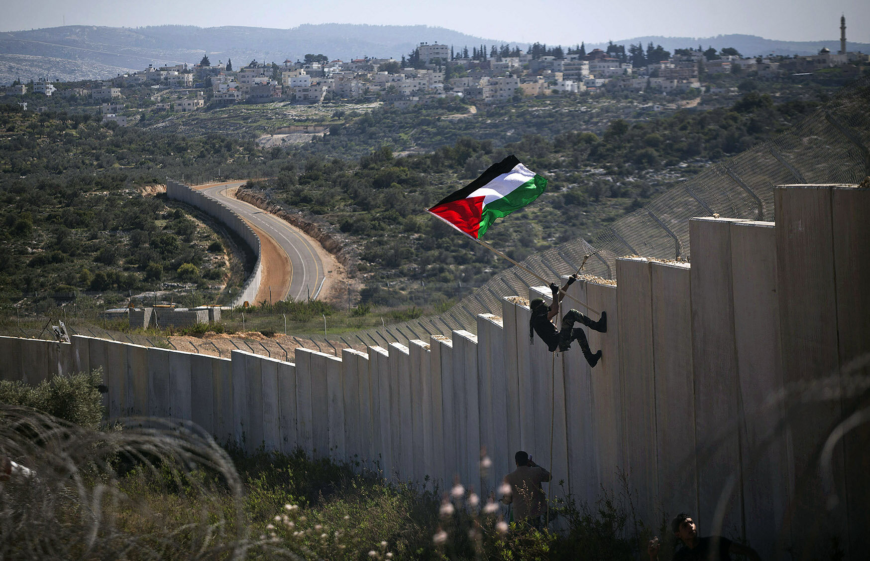 El muro de separación de Israel en Bil'in, donde un manifestante palestino planta una bandera, el asentamiento de Modi'in Illit al fondo (foto por cortesía de Oren Ziv, el Proyecto GroundTruth ).
