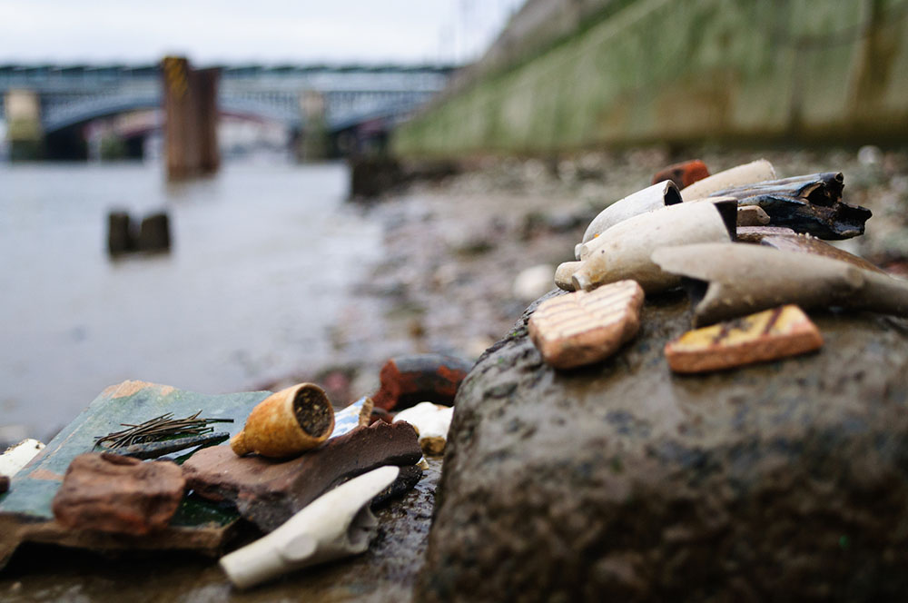Mudlark findings- close up of broken clay pipes, pottery shards and other fragments found on the River Thames while mudlarking, with the river and bridge in the background 