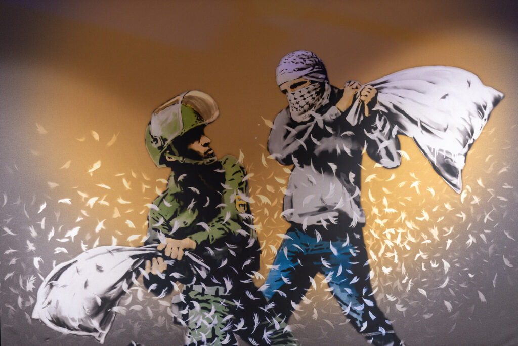 Banksy, Pillow Fight, 2017, Palestinian rebel and Israeli border police officer do a pillow fight. Artwork, originally done by the anonymous English street graffiti artist Banksy with political message showed in exhibition - Banksy a Genius Mind - in Muelheim, Germany. Banksy hoped, that Israelis and Palestinians started much needed dialogue so that a conflict doesn t require heavy artillery.